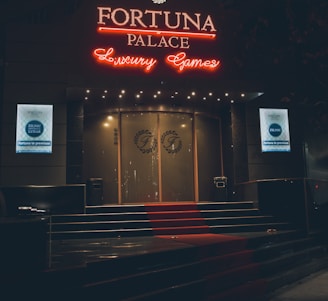 A neon-lit casino entrance with a red carpet leading up to large double doors. The sign reads 'Fortuna Palace Luxury Games' in bright red and white neon lights. The dimly lit atmosphere suggests a night-time setting. There are two digital displays on either side of the entrance showing promotional content.
